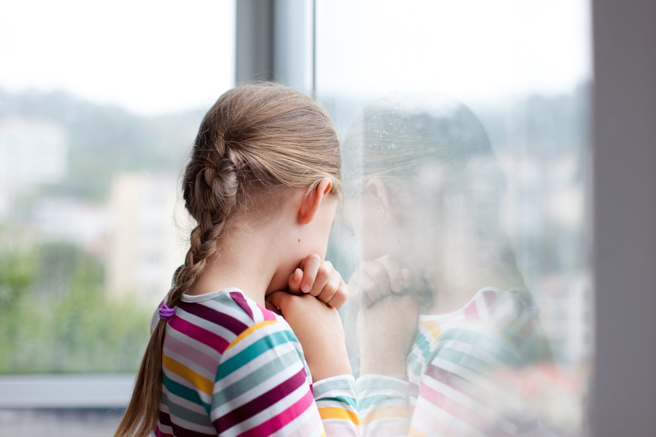 Sad child looking through rainy window at home. Upset kid in self isolation during quarantine.